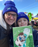 A woman wearing glasses, a blue school cap, and a jacket poses next to a child, who is also wearing the blue school cap, and is holding up a piece of artwork.
